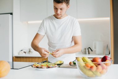 Ev yemeği konsepti. Adam tahıllı ekmek, domates ve peynirden yapılmış sandviçe avokado dilimleri koyuyor. Modern beyaz mutfakta avokadoyla tost yapma süreci.