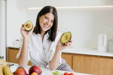 Mükemmel olgunlaşmış avokadonun yarısını elinde gösteren ve modern beyaz mutfakta taze meyve ve sebzelerin arka planında gülümseyen genç ve mutlu bir kadın. Sağlıklı gıda konsepti. Ev yemeği.