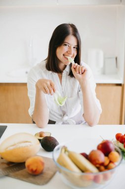 Sağlıklı gıda konsepti. Genç mutlu kadın yeşil marul yaprağı yiyor ve modern beyaz mutfakta taze meyve ve sebzelerin arka planında gülümsüyor. Ev yemeği.