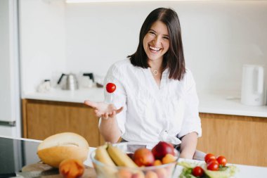 Sağlıklı gıda konsepti. Genç, mutlu bir kadın kiraz domatesi fırlatıyor ve modern beyaz mutfağın arka planında gülümsüyor. Ev yemeği, salata malzemeleri.