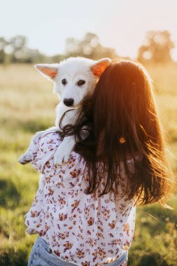 Tarz sahibi genç bir kadın yaz otlağında gün batımında sevimli beyaz bir köpeğe sarılıyor. Mutlu kız sevimli pofuduk köpek yavrusunu tutuyor. Güzel atmosferik bir an. Evlat edinme kavramı.
