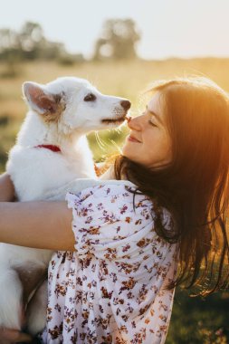 Şirin pofuduk yavru köpek öpen kız. Güzel atmosferik bir an. Tarz sahibi genç bir kadın yaz otlağında gün batımında sevimli beyaz bir köpeğe sarılıyor. Evlat edinme kavramı