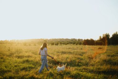 Havalı hippi kız yaz otlağında şirin beyaz bir köpek yavrusuyla yürüyor. Ilık gün ışığında köpeğiyle yürüyüşe çıkmış genç bir kadın. Evlat edinme konsepti, sadık dostum.