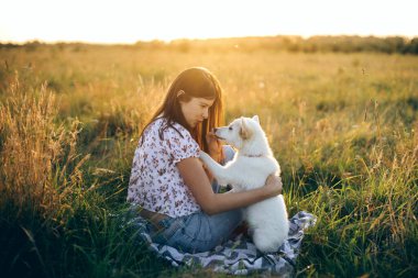 Mutlu bir kadın ve şirin beyaz bir köpek yavrusu gün batımında yaz otlaklarında piknik yapıyorlar. Şık kız, sevimli tüylü köpeğiyle rahatlıyor. Sadık bir dost. Hayvanla yaz seyahati