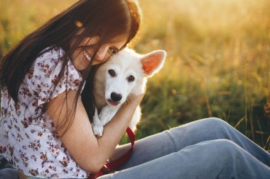 Tarz sahibi kız, sevimli köpeğiyle piknikte rahatlıyor. Evcil hayvanla yaz tatili. Mutlu kadın gün batımında yaz çayırında sevimli beyaz köpek yavrusuna sarılıyor. Otantik güzel an.