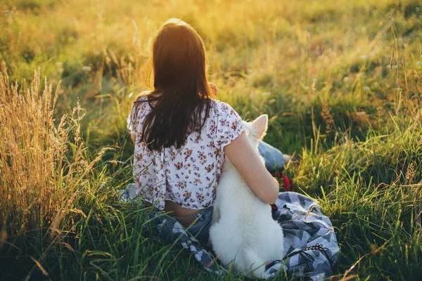 一个女人和可爱的白狗坐在一起 看着夏日的草地上的落日 在温暖的夕阳西下 一个时髦的女孩和她可爱的毛茸茸的小狗在一起放松 忠诚的朋友与宠物的暑期旅行 — 图库照片