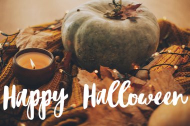 Mutlu Cadılar Bayramı tebrik kartları. Balkabağı, sonbahar yaprakları, mum, sarı örülmüş süveter üzerine sıcak ışıklar üzerine el yazısı Mutlu Cadılar Bayramı metni