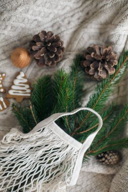 Yeşil ladin dallı ağ pamuk torbası, zencefilli noel kurabiyesi ve beyaz örgü süveterin üzerinde çam kozalağı. Yeniden kullanılabilir alışveriş çantası, ahşap kış süslemeleri, plastik ücretsiz tatiller.