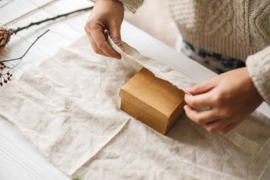 Plastiksiz Noel hediyesi, sıfır ziyan tatili. Samimi süveter giymiş kadın elleri, Noel hediyesini kırsal ahşap arka planda keten kumaşla sarıyorlar..