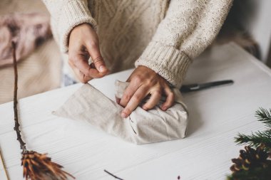 Kadın eli, keten kumaşla yeşil dallı, kozalaklı, makaslı ahşap masaya Noel hediyesini sarıyordu. Sade süveterli bir kadın plastik Noel hediyesi hazırlıyor, sıfır ziyan tatili