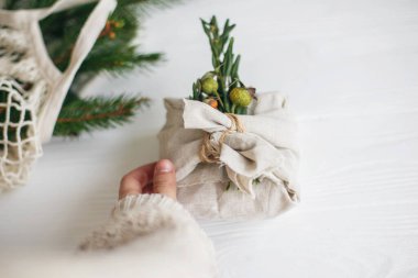 Hand holding stylish christmas gift wrapped in linen fabric with green  branch on rustic white wooden background. Zero waste christmas holidays. Sustainable lifestyle clipart