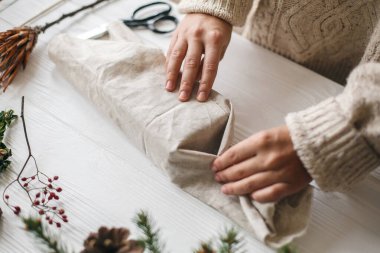 Plastiksiz Noel hediyesi, sıfır ziyan tatili. Kadın elleri rahat süveter içinde keten kumaşla yeşil dallı, çam kozalaklı, makaslı ahşap masaya Noel hediyesini sarıyorlar..