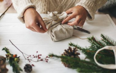Kadın eli, keten kumaşla yeşil dallı, çam kozalaklı, makaslı ve sicimli beyaz kırsal masa üzerinde şık bir Noel hediyesi paketliyor. Sade süveterli bir kadın plastik Noel hediyesi hazırlıyor.