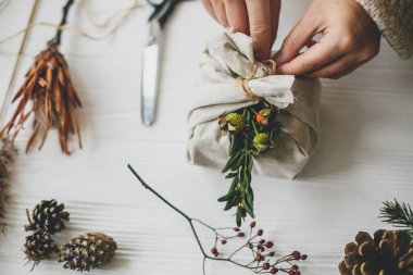 Kadın sıfır atık noel hediyesi hazırlıyor. Beyaz kır masasında doğal otlar ve çam kozalakları ile yeşil dallı keten kumaşlarla süslenmiş eller..