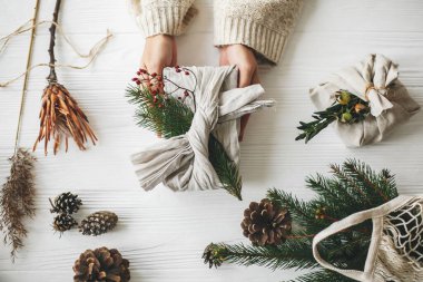 Yeşil köknar dalıyla keten kumaşa sarılı zarif Noel hediyesini tutan eller ve çam kozalaklı kırmızı böğürtlenler. Sıfır kayıp Noel, plastiksiz tatil