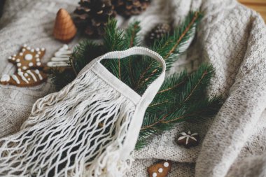 Yeniden kullanılabilir alışveriş çantası, ahşap kış süslemeleri, plastiksiz tatiller. Yeşil ladin dallı ağ pamuk çuvalı, zencefilli noel kurabiyeleri ve beyaz örülmüş süveterin üzerindeki çam kozalakları..