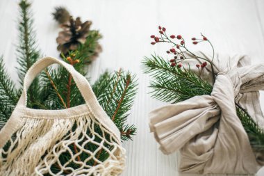 Yeşil dallı keten kumaşa sarılı şık bir Noel hediyesi ve kırsal ahşap arka planda yeşil ladin ile yeniden kullanılabilir bir alışveriş çantası. Sıfır noel ziyanı. Sürdürülebilir yaşam tarzı