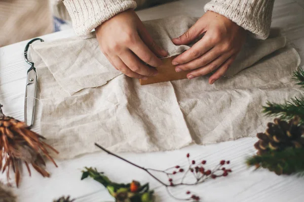 Kadın elleri rahat süveter içinde keten kumaşla yeşil dallı, çam kozalaklı, makaslı ahşap masaya Noel hediyesi sarıyorlar. Plastiksiz Noel hediyesi, sıfır ziyan tatili..