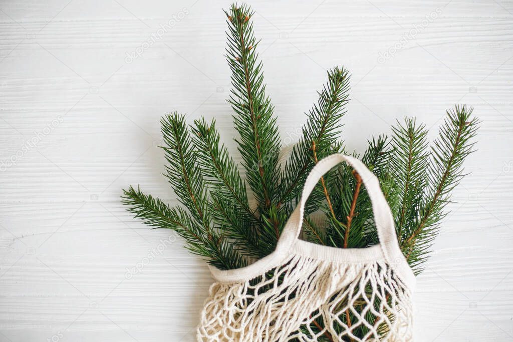 Net shopping bag with winter decorations, zero waste holidays. Sustainable lifestyle. Reusable cotton bag with green spruce branches on white rustic background.