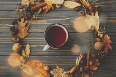 Merhaba sonbahar! Kırsal sonbahar düzlüğü. Yapraklardan, meyvelerden, meşe palamudlarından ve koyu ahşap arka planda koyu renkli çam kozalaklarından yapılmış çay ve sonbahar çelenkleriyle klasik metal bardak..