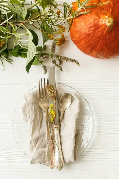 Eco friendly Thanksgiving feast. Stylish plate with cutlery and autumn decorations, pumpkin, natural branches and autumnal flowers on white table. Rustic table setting decor