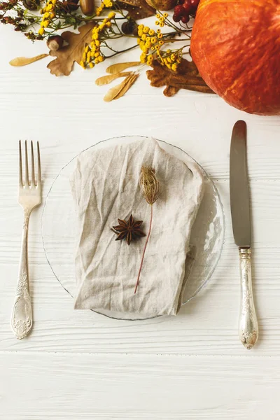 Eco friendly Thanksgiving feast. Stylish plate with cutlery, linen napkin, anise and autumn leaves, pumpkin,  autumnal flowers on white table. Rustic table setting decor