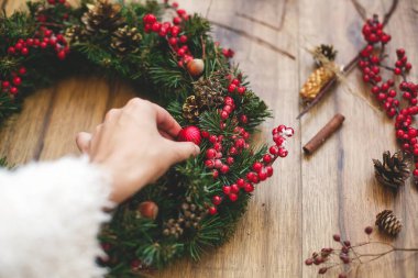 Kırsal ahşap arka planda geleneksel Noel çelengi yapan kişi. Çiçekçi el süsü, pamuk, kırmızı böğürtlen ve çam kozalaklarıyla Noel çelengi süslüyor. Tatil atölyesi