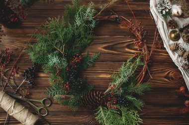 Ahşap masa üzerinde kırsal noel çelengi. Kırmızı böğürtlenli modern noel çelengi, yeşil sedir dalları ve kır masasında çam kozalağı makas, sicim ve şenlikli dekorasyonla.