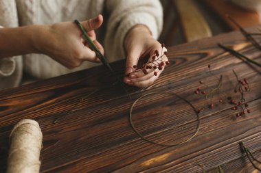 Kırsal ahşap masada Noel çelengi yapmak, bayram gelişi. Çiçekçi elinde makasla kırmızı böğürtlenli çelenk yapıyor. Şenlik Atölyesi