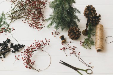 Beyaz ahşap masada rustik noel çelengi ayrıntıları. Kırmızı böğürtlen, iplik, makas, çam kozalağı ve kırsal arkaplanda mum