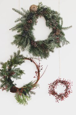 Beyaz duvarda Rustik Noel çelengi asılı, şenlikli dekorasyon. Kırmızı böğürtlenli ve köknar dallı yaratıcı doğal ve farklı noel çelenkleri, beyaza izole edilmiş. Mutlu Noeller.