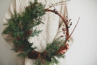 Beyaz arka planda kadın ellerinde Rustik Noel çelengi. Modern noel çelengi taşıyan şık örülmüş süveterli kadın köknar dalları, böğürtlenler ve koni, bayram gelişimi.