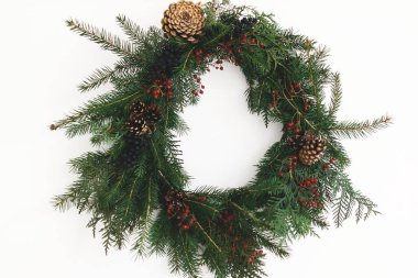 Beyaz duvarda asılı Rustik Noel çelengi, şenlikli modern dekorasyon. Kırmızı böğürtlenli ve köknar dallı yaratıcı bir Noel çelengi, beyazla izole edilmiş. Mutlu Noeller.