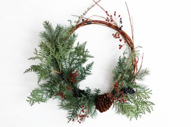 Beyaz duvarda asılı Rustik Noel çelengi, bayram dekorasyonu. Kırmızı böğürtlenli ve köknar dallı yaratıcı modern noel çelengi, beyazla izole edilmiş. Mutlu Noeller.