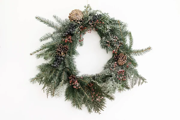 Beyaz duvarda asılı Rustik Noel çelengi, şenlikli modern dekorasyon. Kırmızı böğürtlenli ve köknar dallı yaratıcı bir Noel çelengi, beyazla izole edilmiş. Mutlu Noeller.