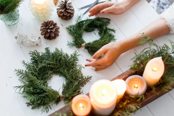 Rustic christmas wreath with candles, pine cones, scissors and ornaments on white wooden table. Female hands making simple stylish christmas wreath with fir branches, holiday advent