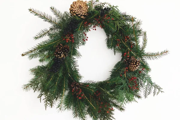 Beyaz duvarda asılı Rustik Noel çelengi, şenlikli modern dekorasyon. Kırmızı böğürtlenli ve köknar dallı yaratıcı bir Noel çelengi, beyazla izole edilmiş. Mutlu Noeller.