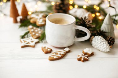 Merhaba kış, sıcacık karamsar görüntü. Ev yapımı zencefilli kurabiye, çam kozalağı süslemeleri, köknar dalları ve beyaz ahşap masa üzerindeki sıcak ışıklarla süslü şık beyaz bardakta sıcak kahve.