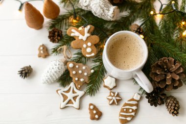 Ev yapımı zencefilli kurabiye, çam kozalağı süslemeleri, köknar dalları ve beyaz ahşap masanın üzerindeki sıcak ışıklarla süslü şık beyaz bardakta sıcak kahve. Merhaba kış, samimi karamsar görüntü.