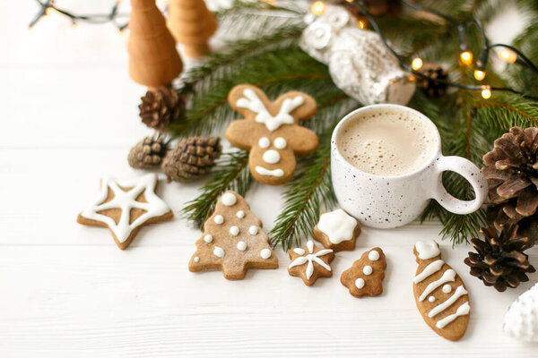 Горячий кофе в стильной белой чашке с домашним пряничным печеньем и сосновыми шишками украшения, ель ветви и теплые огни на белом деревянном столе. Адская зима, уютный капризный образ