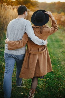 Mutlu şık bir çift, sıcak günbatımı ışığında sonbahar çayırlarında sarılıp yürüyorlar. Romantik duygusal an. Genç, şık kadın ve erkek sonbahar tarlasında dinleniyor.
