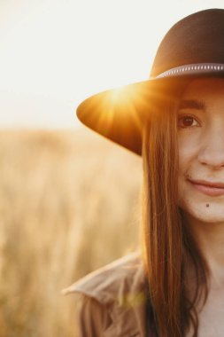 Sonbahar tarlasında gün batımında sakin bir kadının portresi. Şapkalı genç, şık bir bayan otların arkasında poz veriyor ve güneş ışığı altında otlar. Duygusal an.