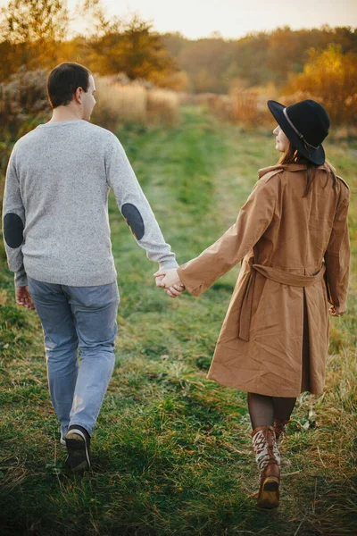 暖かい日没の光の中で秋の牧草地で手を取り合って歩く幸せなスタイリッシュなカップル ロマンチックな官能的な瞬間 若いファッショナブルな女性と男は秋のフィールドでリラックス — ストック写真