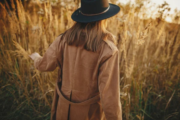 暖かい日没の秋のフィールドで草の間を歩くスタイリッシュなBohoの女性 若いファッショナブルな女性が茶色のコートとハーブでリラックス帽子 バックビュー ロマンチックな本物の瞬間 — ストック写真