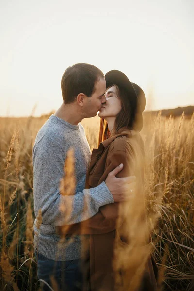 秋のフィールドで暖かい夕日の光を受け入れるスタイリッシュなカップル 若いファッショナブルな女性と男は太陽の下で草やハーブの間で抱き合ってキスをします ロマンチックな本物の瞬間 — ストック写真