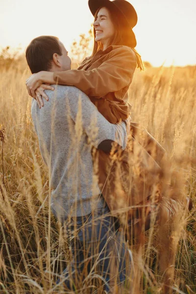 秋のフィールドで暖かい夕日の光の中で踊るスタイリッシュな幸せなカップル 太陽の下で草やハーブの間で若いファッショナブルな女性と男が渦巻く ロマンチックな本物の瞬間 — ストック写真