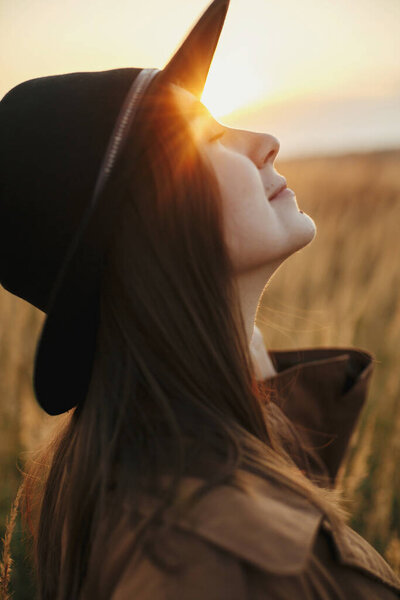 Portrait of calm boho woman in warm sunset light in autumn field. Young stylish female in hat posing on background of grass and herbs in sunshine. Sensual moment