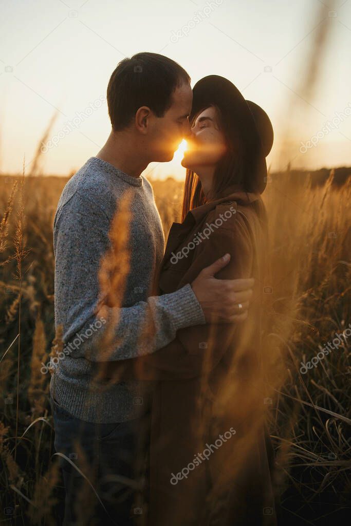 Stylish sensual couple embracing in warm sunset light in autumn field. Young fashionable woman and man gently hugging among grass and herbs in sunshine. Romantic authentic moment