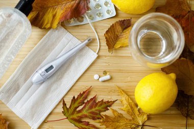 Sonbahar gribi ya da koronavirüs mevsiminde bağışıklık sistemini güçlendirin. D vitamini, C vitamini ve çinko hapları maske, dezenfeksiyon jeli, limon, termometre ve dökülen yapraklarla ahşap masada.