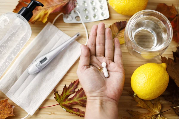 Hand with vitamin D, vitamin C and zinc pills on background of wooden table with fall leaves, face mask, sanitizer, lemons. Boost immune system, coronavirus treatment. Stay healthy